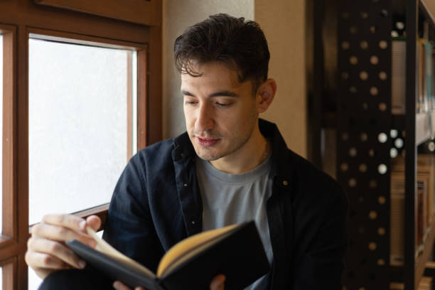 istock Young man reading a book by the window in a library 2193281550