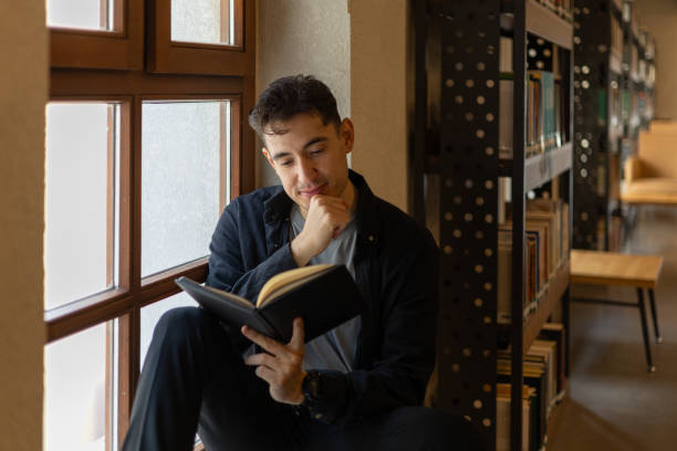 istock Young man reading a book in a library by the window 2193281570