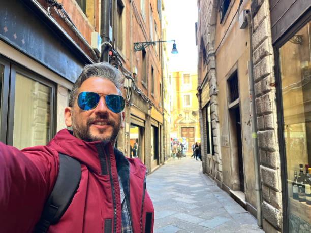 Latin male tourist wearing sunglasses and a maroon jacket, capturing a selfie in a narrow street of Venice, Italy, during the vibrant Carnival festivities, surrounded by colorful architecture