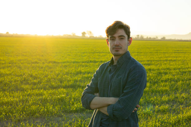 Confident young farmer with arms crossed, standing proudly in a cultivated wheat field at sunset, gazing directly at the camera while surrounded by the beauty of nature