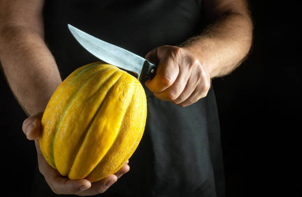 istock A skilled hand holds a bright yellow melon, poised with a gleaming knife ready to slice. The contrasting colors highlight the freshness and texture of the fruit against a dramatic black background 2193299599