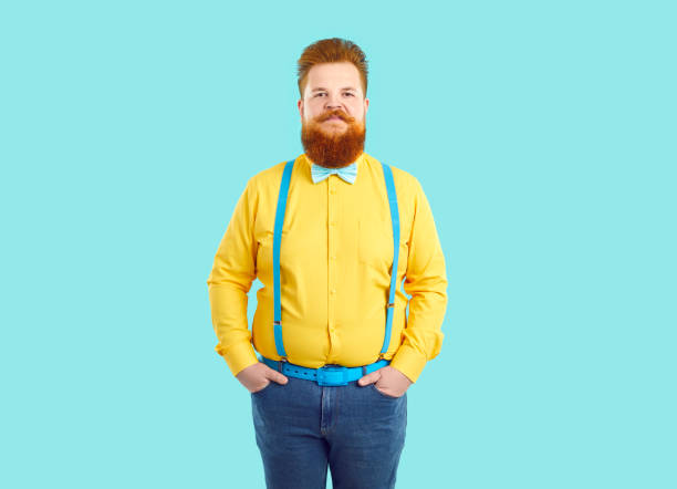 Portrait of extravagant confident and stylish chubby funny man on light blue background. Serious red-haired fat man with mustache and beard wearing jeans, yellow shirt, blue suspenders and bow tie.