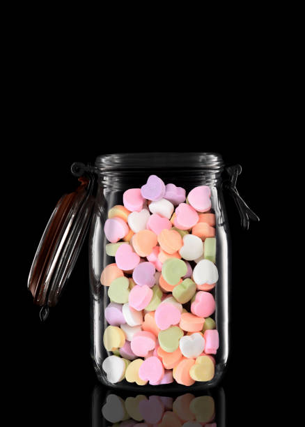 A glass storage or canning jar filled with Valentines Candy Hearts isolated on black with reflection, with lid open.