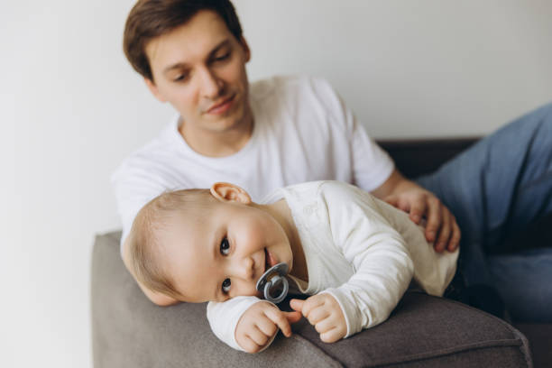 istock Baby lying on sofa with pacifier and looking at camera, father watching 2193317310