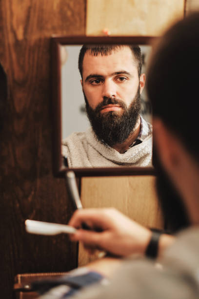 istock Young and handsome man with a beard holds a Straight Razor in his hand for shaving in front of a mirror. 2193324982