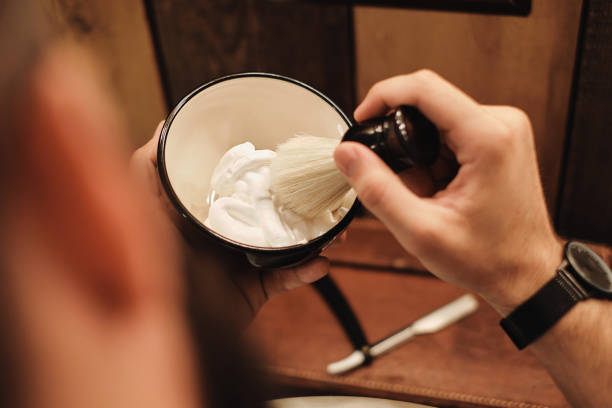 A young and handsome man with a beard is preparing shaving cream with a brush in a bowl. In front of a wooden wall.