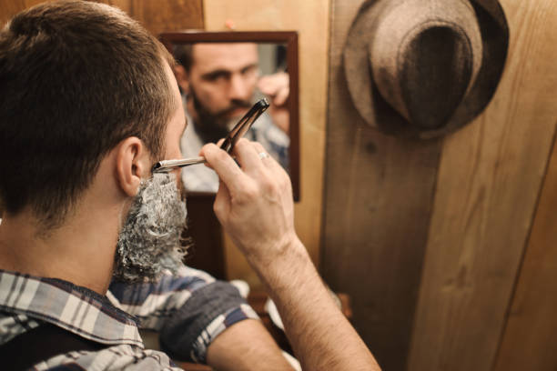 A young and handsome man with a beard begins to shave in front of a mirror. A wooden wall in front of.