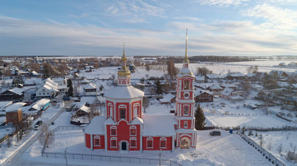 Red church with a white roof is surrounded by snow. The snow is piled up on the roof and the ground. The church is surrounded by houses and buildings. The sky is cloudy. The ancient Russian city of Suzdal