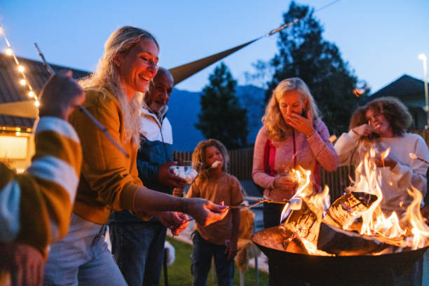 A cheerful, diverse, multigenerational group seen engaging in an outdoor camping activity, roasting marshmallows over an open fire during twilight. Individuals of different ages share a joyful moment together.