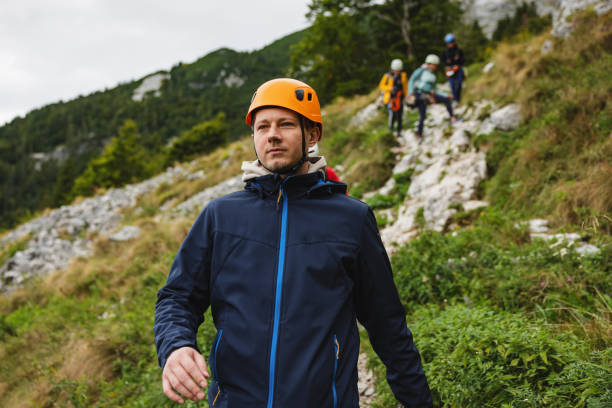 A group of diverse friends prepare for a ziplining activity in a mountainous area. The male with a helmet in the foreground is followed by others equipped with safety gear.