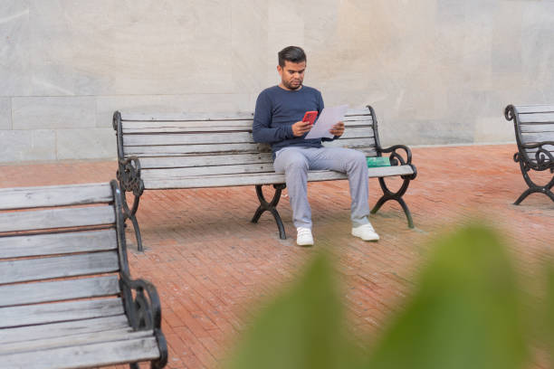 Focused young businessman working remotely, checking his phone and reading a document while sitting on a bench