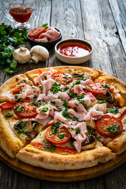 istock Pizza with ham, mozzarella cheese, sliced tomatoes and white mushrooms on wooden table 2193364814