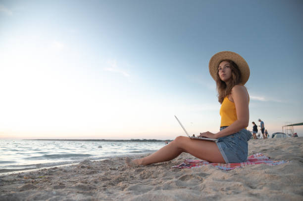 istock Work from anywhere. Side view of young woman freelancer in straw hat working on laptop while sitting on tropical sandy beach at sunset 2193375929