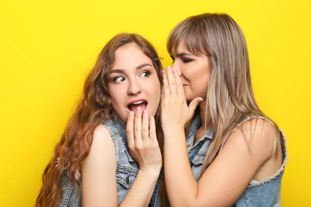 istock Two young woman whispering a secret on yellow background 2193393252