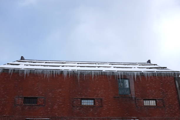 Large icicles hanging from the edge of a roof above a brick wall with two small windows and a vent.