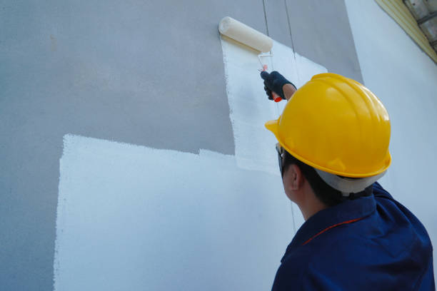 Сonstruction worker is hanging on safety ropes outside a residential building on the ninth floor. He is applying adhesive to the surface of yellow reinforcing mesh, which is being fixed onto polystyrene insulation panels. The worker is insulating the building facade.