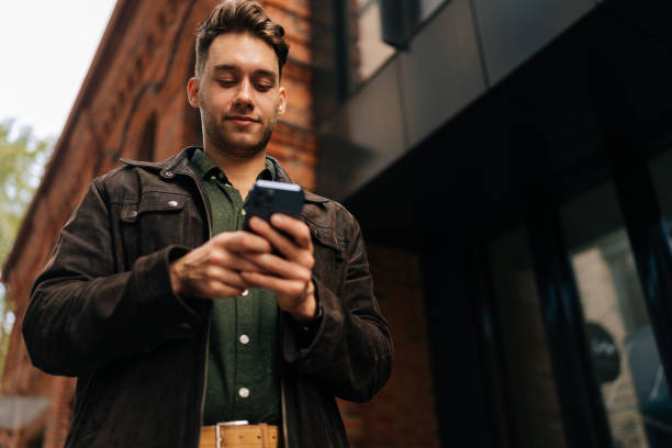 istock Low-angle view of handsome bearded man standing on city street, using smartphone to check messages, browse internet, stay connected enjoying urban environment. 2193433427
