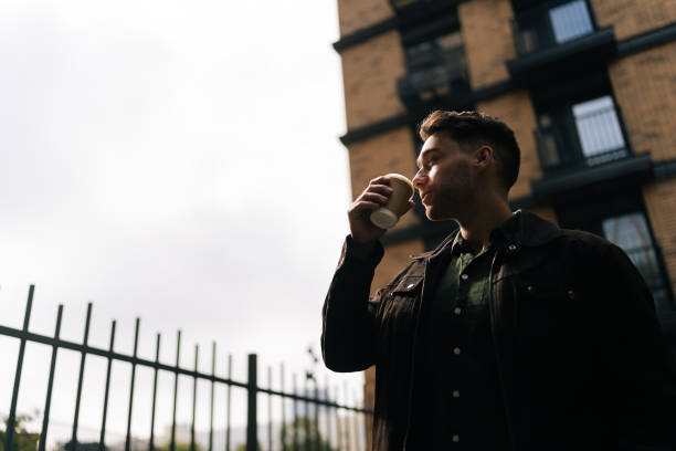 istock Low-angle view of handsome man thoughtful looking away enjoying drinking coffee standing in urban alleyway by brick buildings, embodying casual city lifestyle 2193433435