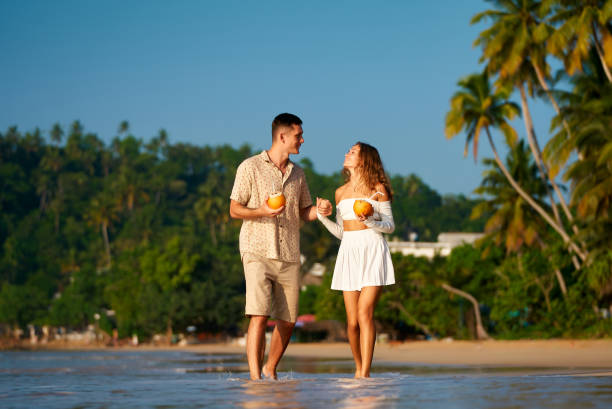 istock Couple stroll barefoot on sandy beach, holding coconuts, sipping drinks, enjoying sunset. Casual date, tropical vacation vibe. Smiles, flirty conversation, ocean backdrop, intimate walk together. 2193433798