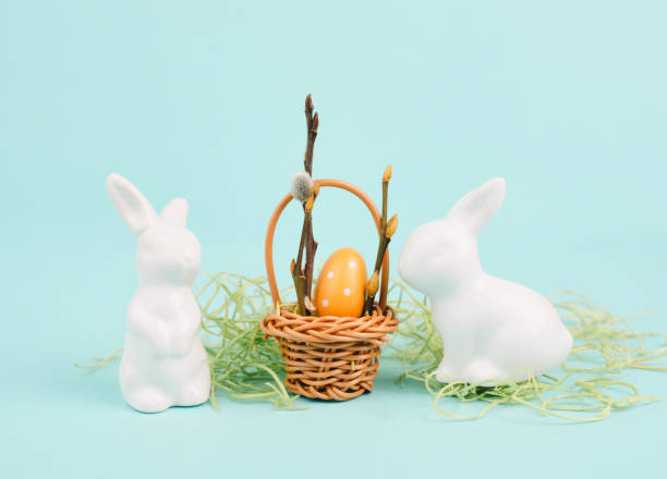 istock Easter rabbit or bunny sitting on green grass, wicker basket with colored egg, pastel blue background, holiday greeting card 2193434064