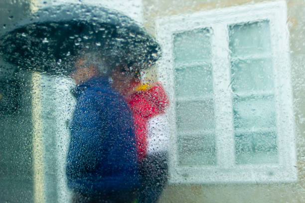 Defocused view of couple with umbrella walking along the sidewalk, city street , raindrops on a glass  in the foreground, closed window in the background. Galicia, Spain.