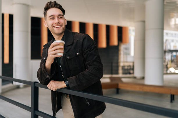istock Cheerful young man relaxing during coffee break, leaning on railing standing by modern office building. Happy bearded male wearing casual attire smiling looking away. 2193441825