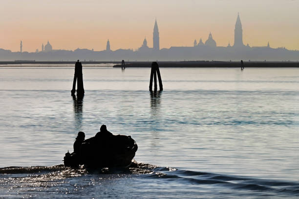 Silhouette of motor boat on calm waters of Venetian lagoon at sunset from Burano island with Venice in the distance