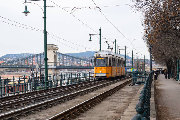 Budapest, Hungary - December 22, 2024: Classic yellow orange colored tram runs along the Danube River in Budapest. The famous chain bridge in the background.