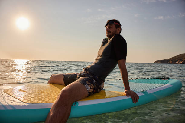 Happy young adult male surfer in a wetsuit sitting on the surfboard and relaxing. It’s a beautiful sunrise.