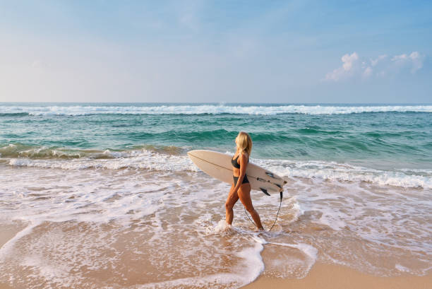 Blonde woman with surfboard walks on sandy beach. Ocean waves crash, sea breeze flows. Active lifestyle and freedom vibe. Surfer girl on sunny day, coastal adventure.