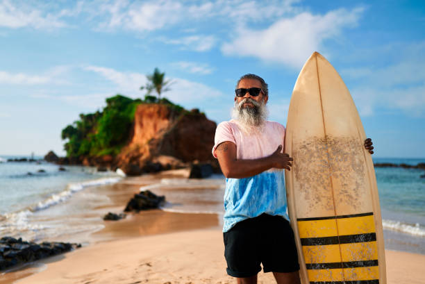 istock Senior man with beard holds surfboard on sunny beach. Elderly surfer smiles, enjoys active retirement on ocean shore. Happy mature male embraces vibrant lifestyle. Summer adventure, freedom. 2193461311