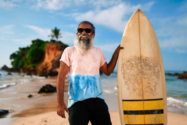 istock Senior man surfer stands on sandy beach with surfboard. Smiling elderly male enjoys active retirement. Ocean waves crash on shore. Surfing in happy golden years. Lifestyle, freedom, adventure. 2193461416