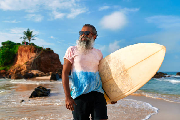 istock Senior black man holds surfboard on sunny beach. Active elderly surfer smiles at ocean waves, enjoying surfing lifestyle. Aged male embraces adventure, healthy living by sea, showing vibrant spirit. 2193461581