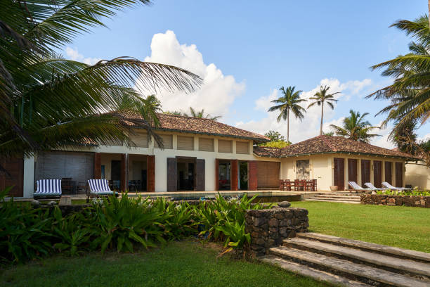 istock Vintage tropical villa with palm trees, stone steps, lush garden. White clouds, sunny sky. Empty sunbeds on lawn. Relaxing vacation retreat. Quiet paradise setting. Architecture blends nature. 2193462442