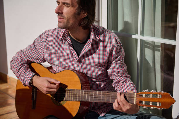 istock Man playing guitar outdoors while seated in sunlight near a window 2193465211