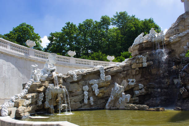 Detailed view of a grand fountain with white sculptures and cascading water, surrounded by greenery and a bright blue sky.