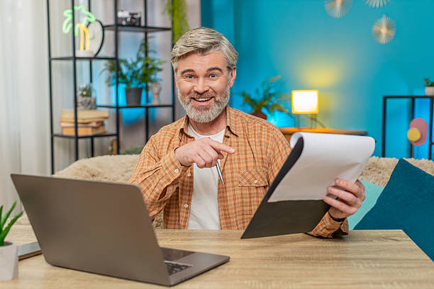 istock Mature man sitting on table with documents and laptop, smiling from successful work at home office 2193470503