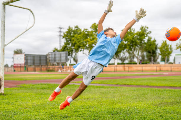 istock Focused soccer goalkeeper leaping mid-air, blocking orange ball during intense training session on grassy field beside running track 2193504497