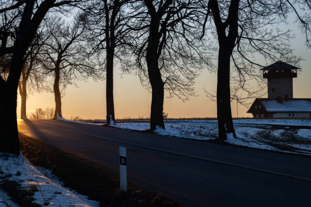 A narrow paved road stretches through open snow-dusted agricultural fields in the Normandy countryside, bordered by distant leafless trees under a calm blue sky.