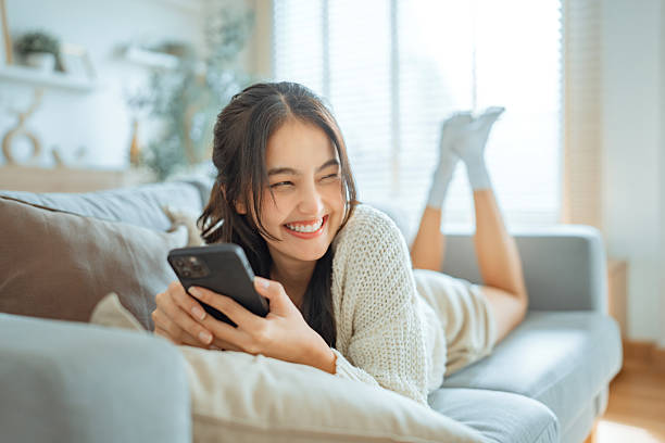 Happy young asian woman lady browsing surfing wireless internet on mobile phone while laying a sofa couch in living room at home apartment, Shopping online via website