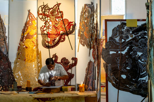 Asian senior man use hammer and chisel to create grand shadow play craft and he concentrate to work in area of workplace with several products are place around workplace area.