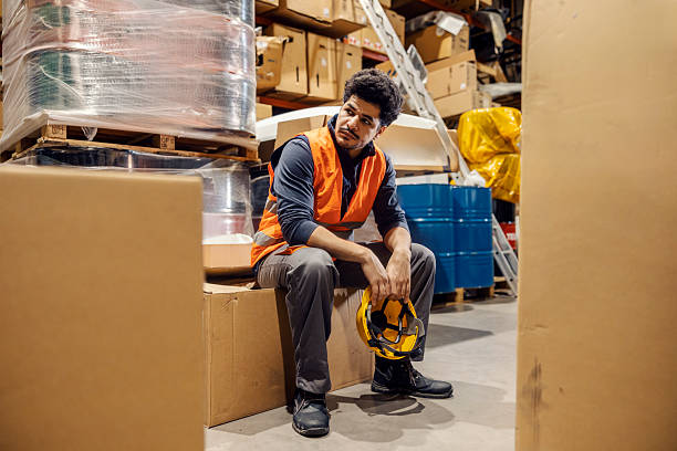 istock Tired interracial warehouse worker with helmet in hands sitting on box surrounded by other boxes. 2193515748