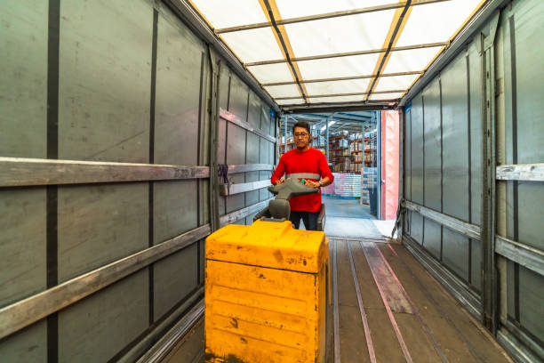 Female worker wearing protective workwear and hardhat is driving a forklift through warehouse with manufacturing materials and boxes.