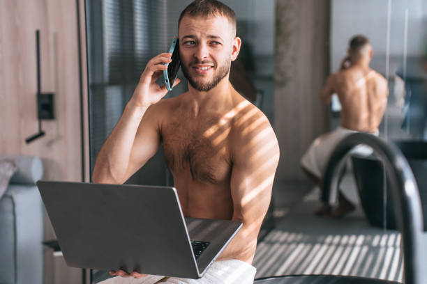 istock A confident man, wrapped in a towel, multitasks with a phone call and laptop amidst gentle morning light, blending relaxation and work. 2193527245