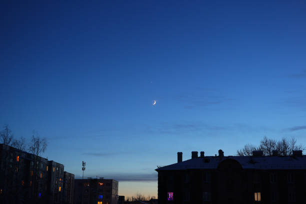 A peaceful evening scene elegantly showcasing a crescent moon set against a beautiful blue twilight sky over rooftops