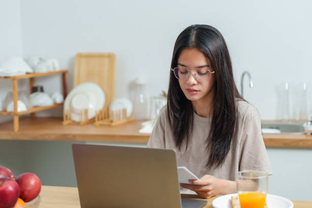 istock Asian woman wearing glasses working on laptop at kitchen table, holding notes, surrounded by modern kitchenware, enjoying productive moment during home relaxation day, cozy and serene. 2193528868