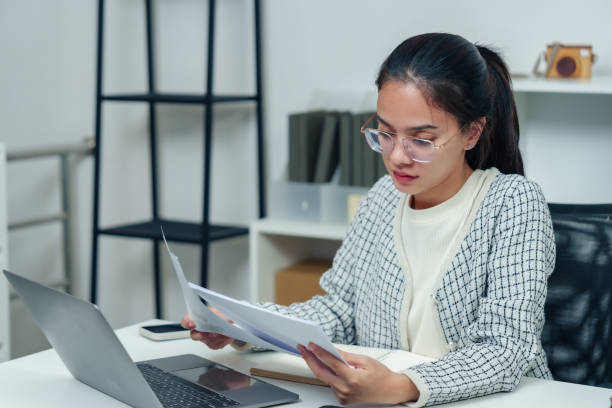 Asian office worker wearing glasses and sweater working on laptop at desk, reviewing documents in modern organized workspace, focusing on professional tasks during office hours trabajador de oficina asiático con gafas y suéter que trabaja en una computadora portátil en el escritorio, revisando documentos en un espacio de trabajo organizado moderno, centrándose en tareas profesionales durante el horario de oficina