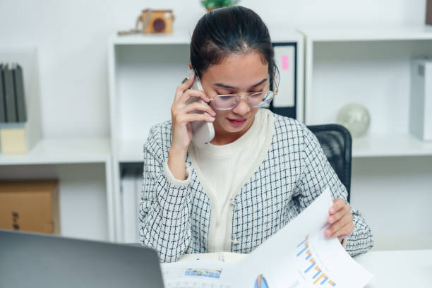 istock Asian office worker reviewing documents while talking on phone, multitasking at organized desk in modern workspace, focusing on professional communication and productivity 2193528971