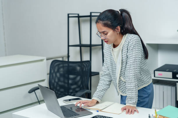 istock Asian office worker leaning over desk using laptop in a modern workspace, wearing professional attire, focusing on productive tasks and work routine in a corporate office setting. 2193528990