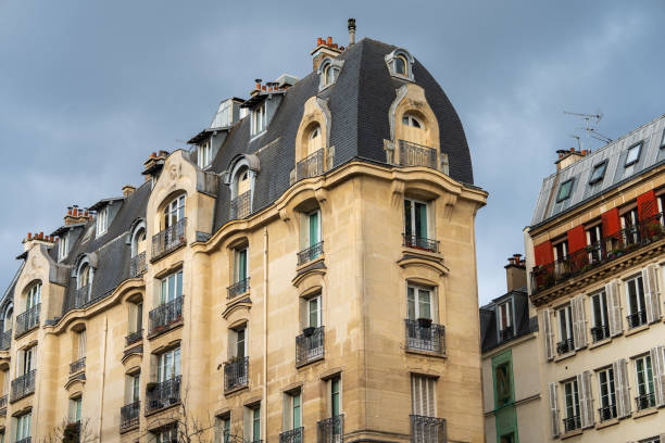 Charming historic buildings rise against a cloudy sky in the heart of Paris. The architectural design showcases intricate details, including ornate balconies and unique roofing styles. The warm tones of the stone façades contrast beautifully with the darker slate roofs. This area reflects the classic Parisian aesthetic, inviting onlookers to appreciate the craftsmanship and history embedded in the structures. The scene captures the essence of urban life in a vibrant city.